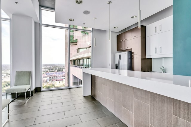 kitchen featuring stainless steel fridge, expansive windows, sink, white cabinetry, and hanging light fixtures