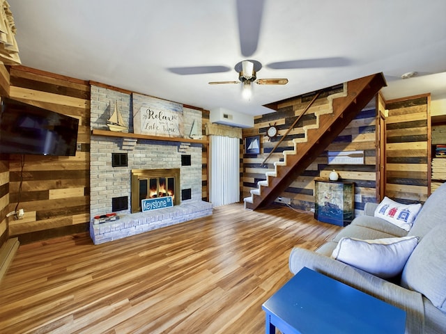 living room featuring wood-type flooring, wooden walls, ceiling fan, and a brick fireplace