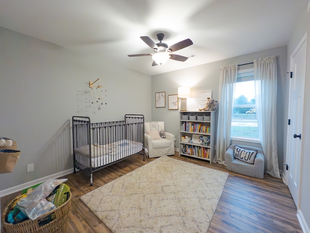 bedroom with a crib, ceiling fan, and dark wood-type flooring