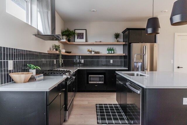 kitchen featuring an island with sink, wall chimney exhaust hood, stainless steel appliances, backsplash, and decorative light fixtures