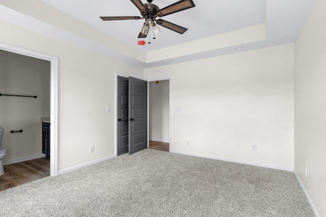 unfurnished bedroom featuring ceiling fan, ensuite bath, a tray ceiling, and carpet