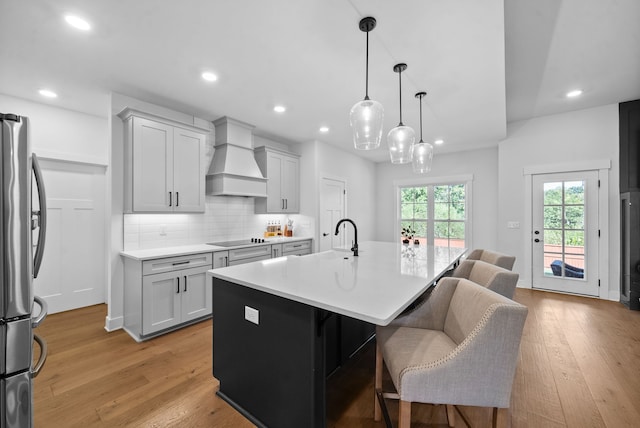 kitchen with hanging light fixtures, stainless steel fridge, a kitchen island with sink, custom range hood, and light wood-type flooring