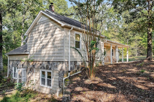 view of home's exterior with a shingled roof, stone siding, a porch, and a chimney