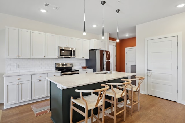 kitchen with pendant lighting, light wood-type flooring, a kitchen island with sink, white cabinets, and appliances with stainless steel finishes