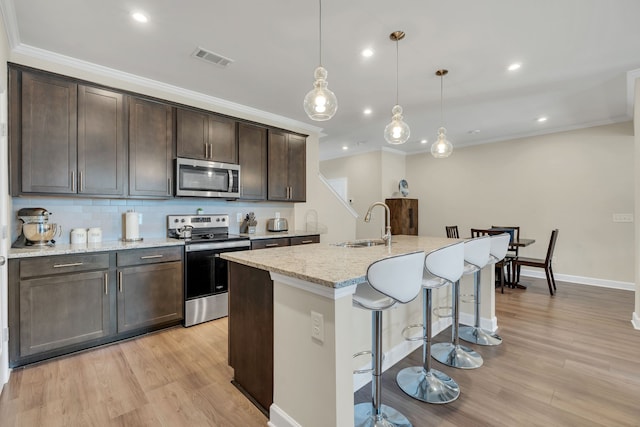 kitchen with light wood-type flooring, light stone counters, an island with sink, hanging light fixtures, and stainless steel appliances