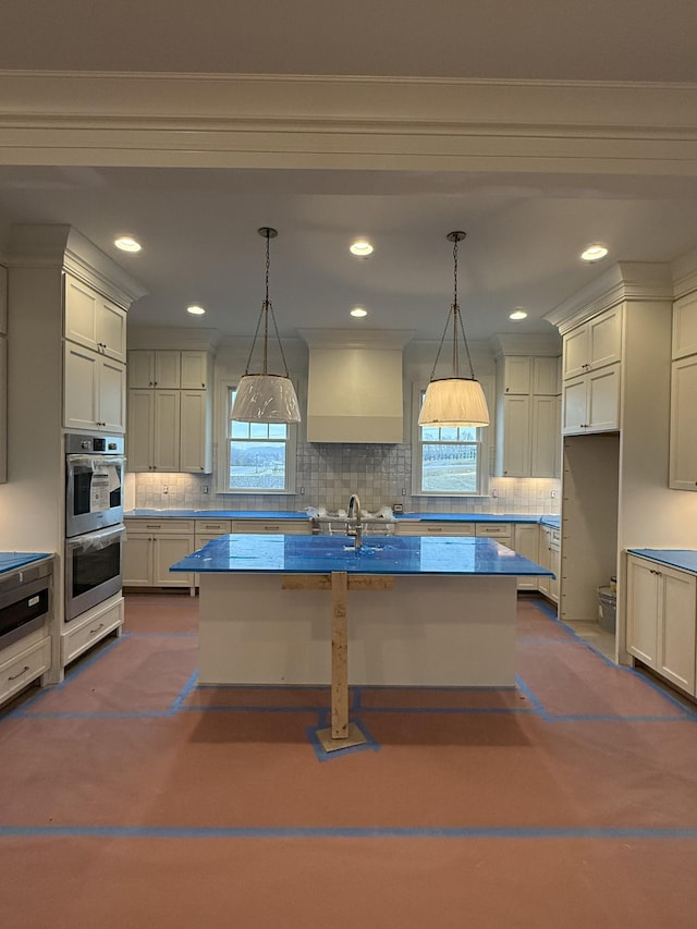 kitchen with an island with sink, stainless steel double oven, custom range hood, and pendant lighting