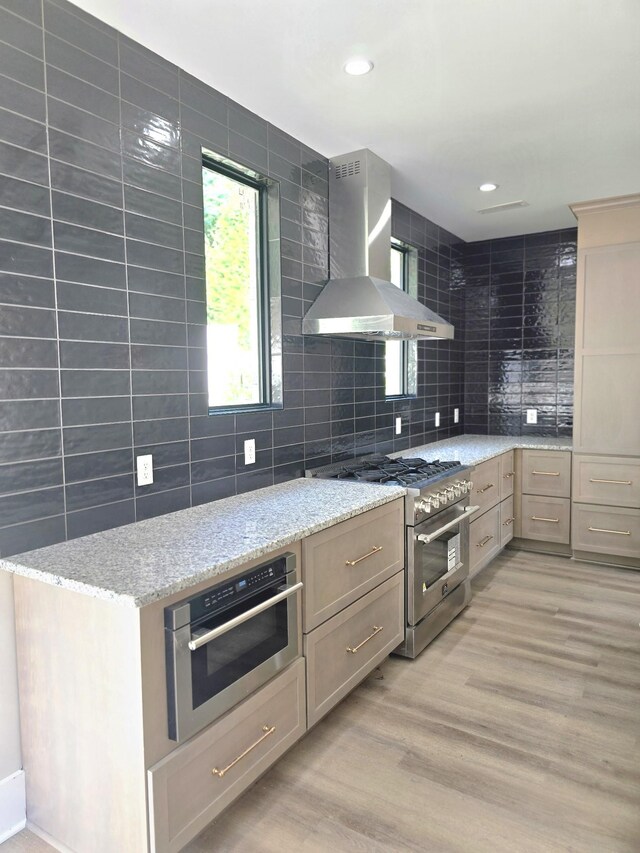 kitchen with wall chimney exhaust hood, light hardwood / wood-style flooring, stainless steel appliances, light brown cabinetry, and light stone countertops