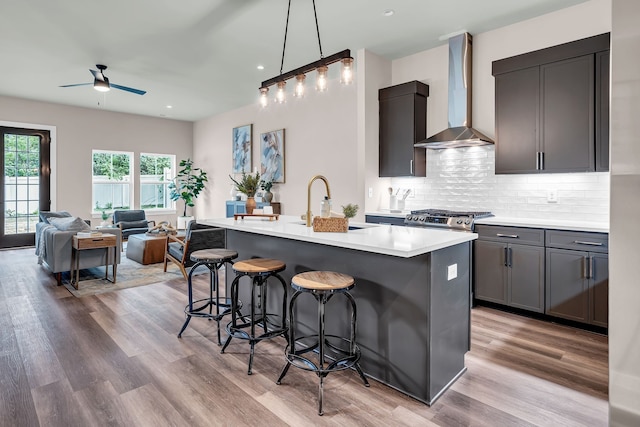 kitchen with ceiling fan, an island with sink, light hardwood / wood-style flooring, and wall chimney range hood