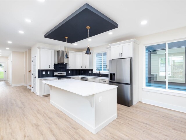 kitchen with light countertops, appliances with stainless steel finishes, white cabinets, a kitchen island, and wall chimney range hood