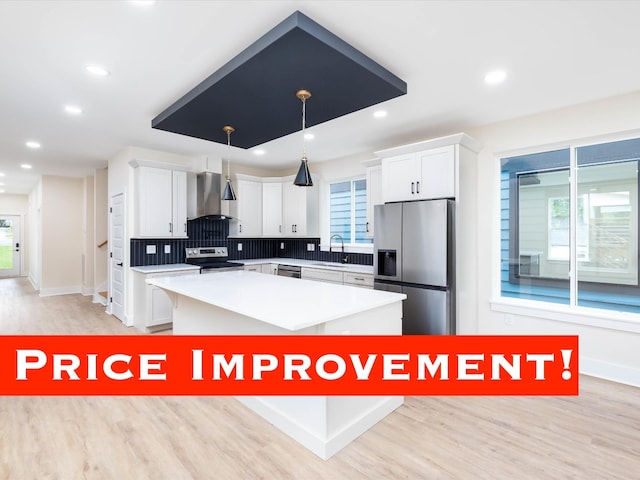 kitchen with white cabinets, appliances with stainless steel finishes, a center island, hanging light fixtures, and wall chimney range hood