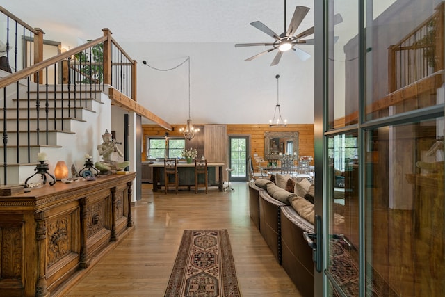 living room featuring ceiling fan with notable chandelier, wood walls, light hardwood / wood-style flooring, and high vaulted ceiling