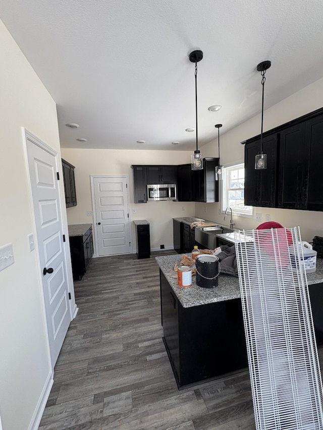 kitchen with dark cabinets, light stone countertops, stainless steel microwave, dark wood finished floors, and pendant lighting