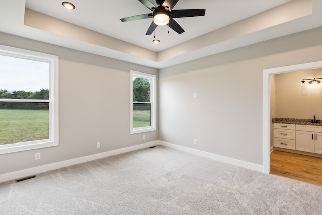 unfurnished bedroom with a raised ceiling, ensuite bath, ceiling fan, and light carpet