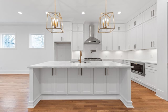 kitchen featuring wall chimney range hood, decorative light fixtures, white cabinets, stainless steel microwave, and an island with sink