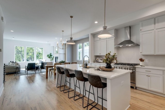 kitchen featuring hanging light fixtures, stainless steel stove, wall chimney exhaust hood, an island with sink, and white cabinetry