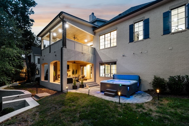 back house at dusk with a lawn, ceiling fan, a balcony, a patio area, and a hot tub