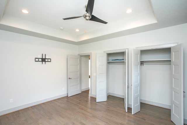 unfurnished bedroom featuring wood-type flooring, two closets, ceiling fan, and a tray ceiling