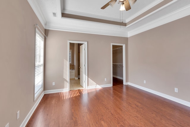 unfurnished bedroom with ceiling fan, a raised ceiling, ornamental molding, wood-type flooring, and a walk in closet