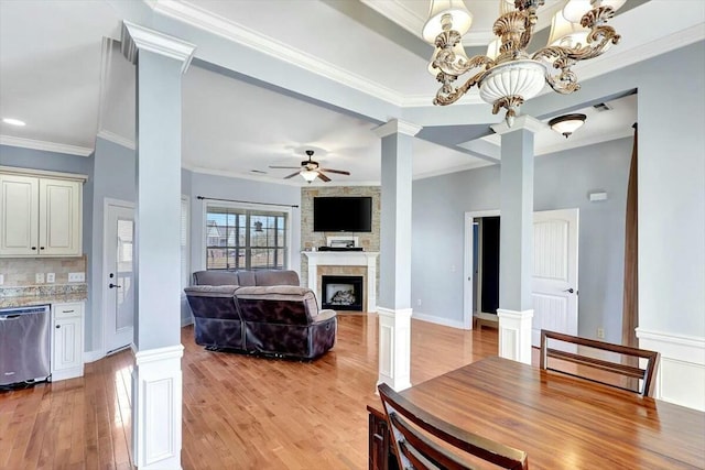 dining space with decorative columns, crown molding, a large fireplace, light wood-style flooring, and ceiling fan with notable chandelier