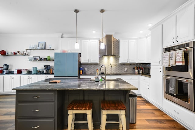 kitchen with stainless steel double oven, white cabinetry, wall chimney range hood, refrigerator, and a kitchen island with sink