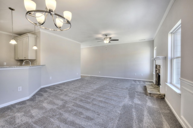 unfurnished living room with sink, ceiling fan with notable chandelier, a stone fireplace, carpet, and crown molding