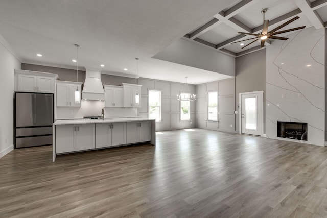 kitchen with custom range hood, stainless steel fridge, white cabinetry, an island with sink, and ceiling fan