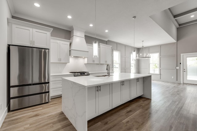 kitchen with appliances with stainless steel finishes, premium range hood, white cabinetry, and a kitchen island with sink