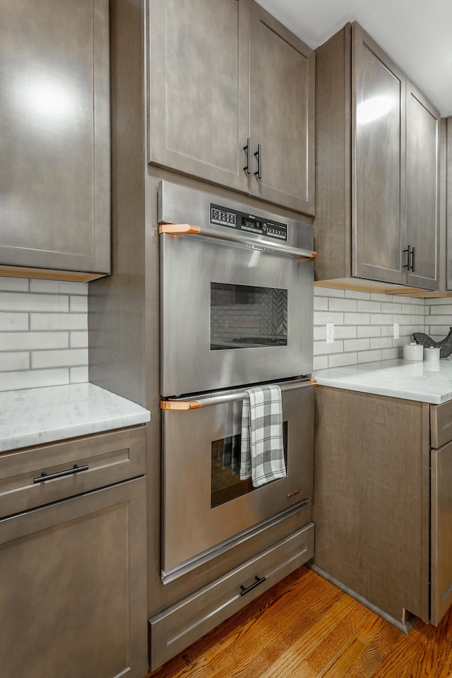 kitchen with light stone countertops, stainless steel double oven, light hardwood / wood-style flooring, and tasteful backsplash