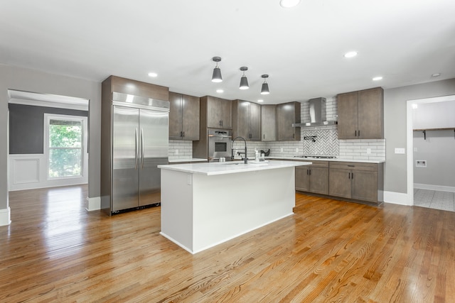 kitchen featuring wall chimney range hood, an island with sink, light hardwood / wood-style floors, dark brown cabinetry, and stainless steel appliances