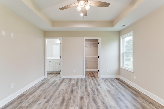 unfurnished bedroom with ensuite bath, ceiling fan, a raised ceiling, a walk in closet, and light wood-type flooring