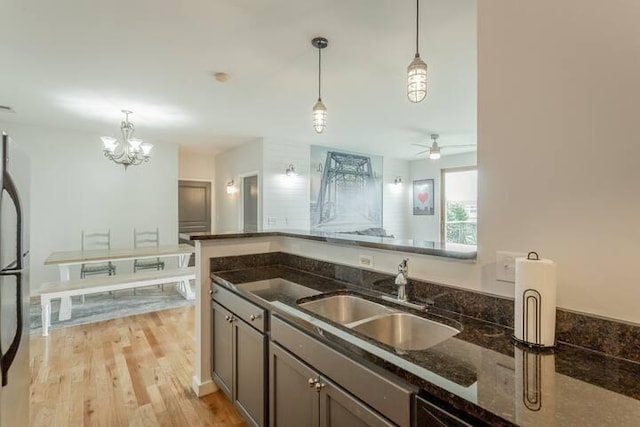 kitchen with pendant lighting, ceiling fan with notable chandelier, sink, light hardwood / wood-style flooring, and dark stone countertops