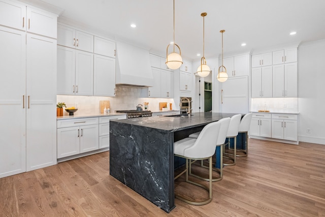 kitchen featuring a kitchen island with sink, sink, white cabinetry, light hardwood / wood-style flooring, and premium range hood