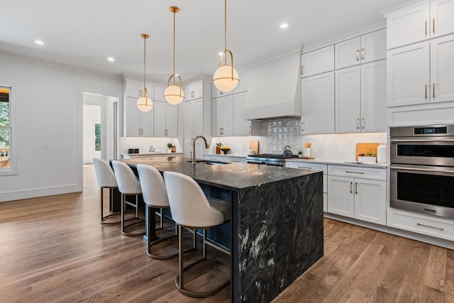 kitchen featuring dark stone counters, sink, an island with sink, white cabinetry, and custom range hood