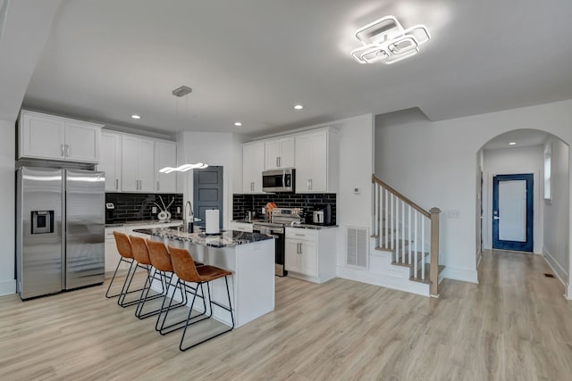 kitchen featuring a kitchen island with sink, light hardwood / wood-style flooring, appliances with stainless steel finishes, dark stone countertops, and decorative backsplash