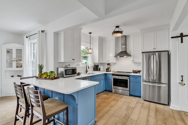 kitchen featuring stainless steel appliances, blue cabinetry, wall chimney exhaust hood, pendant lighting, and a barn door