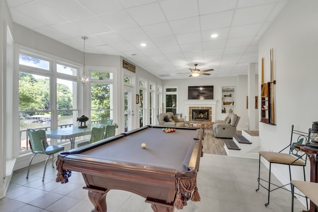 recreation room with light wood-type flooring, ceiling fan with notable chandelier, built in shelves, a brick fireplace, and billiards