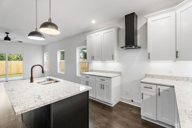 kitchen featuring wall chimney exhaust hood, white cabinets, an island with sink, and light stone counters