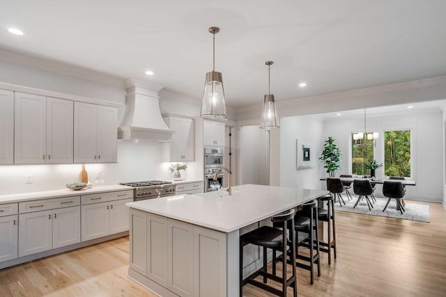 kitchen with custom exhaust hood, decorative light fixtures, a center island with sink, light hardwood / wood-style flooring, and stainless steel appliances