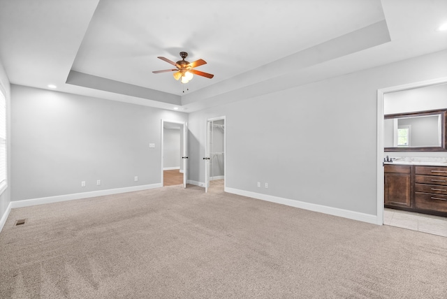 unfurnished bedroom featuring ceiling fan, connected bathroom, a walk in closet, a tray ceiling, and light carpet