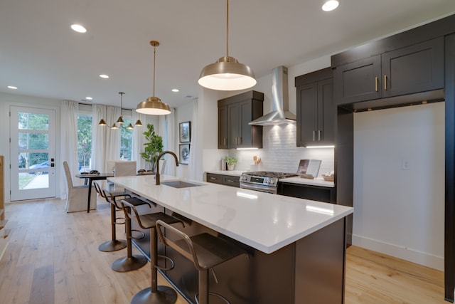 kitchen featuring an island with sink, hanging light fixtures, sink, stainless steel gas range, and wall chimney range hood