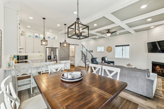 dining room with ceiling fan with notable chandelier, beam ceiling, coffered ceiling, a barn door, and light hardwood / wood-style flooring