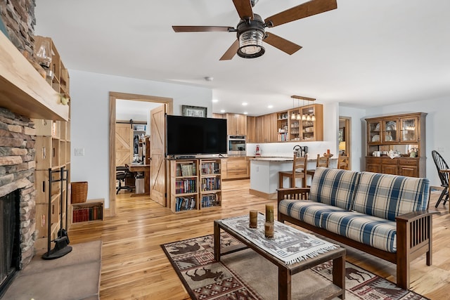 living room with a stone fireplace, light wood-type flooring, ceiling fan, and a barn door