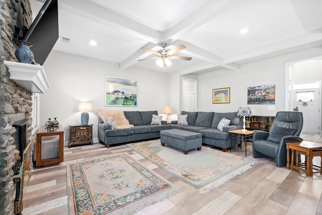 living room with ceiling fan, a stone fireplace, beamed ceiling, and light wood-type flooring