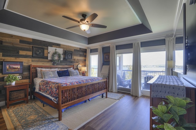 bedroom with ceiling fan, a raised ceiling, wood walls, dark wood-type flooring, and access to exterior