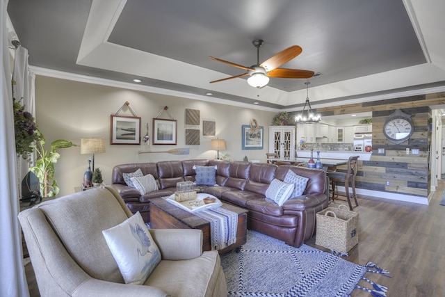 living room featuring wood walls, wood-type flooring, ceiling fan with notable chandelier, and a tray ceiling