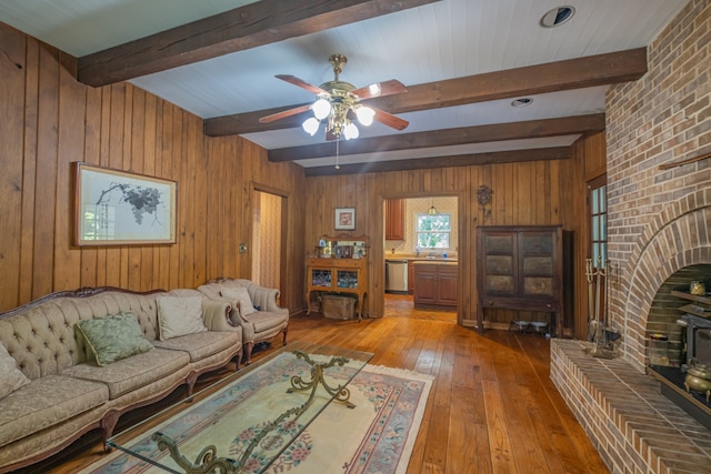 living room featuring ceiling fan, beam ceiling, a wood stove, wooden walls, and hardwood / wood-style floors