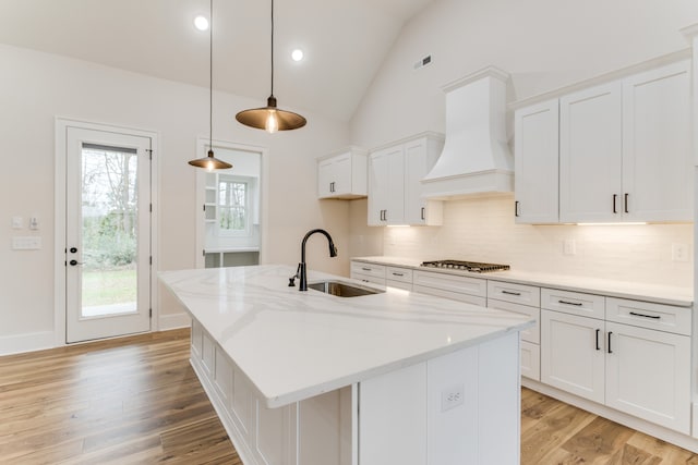 kitchen featuring white cabinetry, sink, premium range hood, an island with sink, and pendant lighting