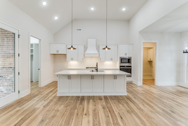 kitchen featuring decorative light fixtures, a kitchen island with sink, appliances with stainless steel finishes, custom range hood, and light wood-type flooring