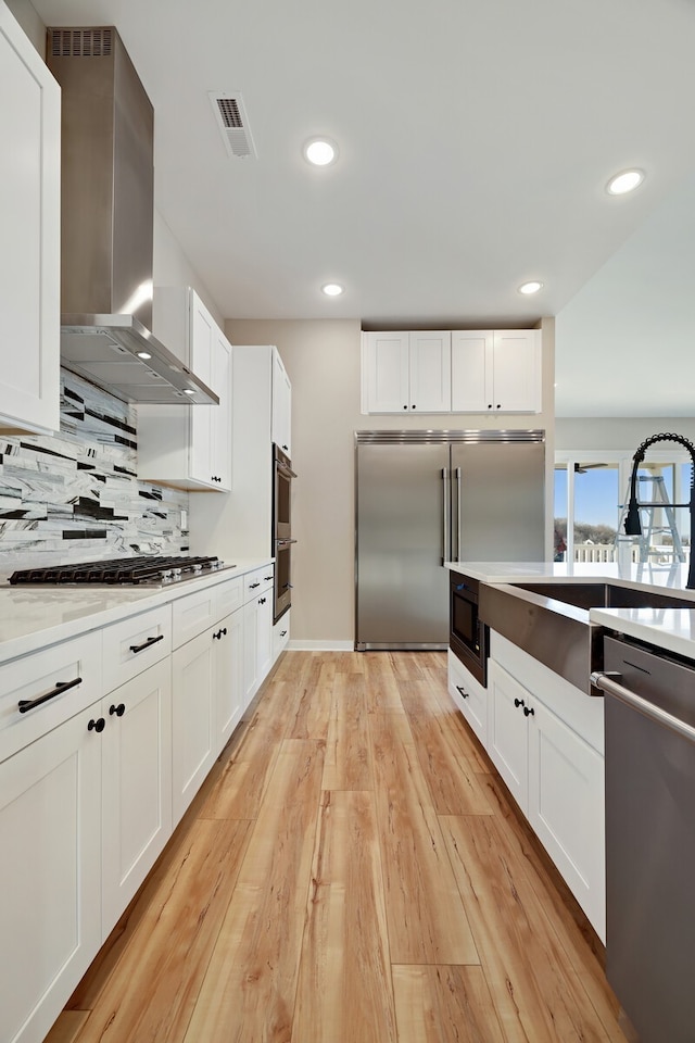 kitchen with light wood-type flooring, white cabinetry, built in appliances, and wall chimney range hood