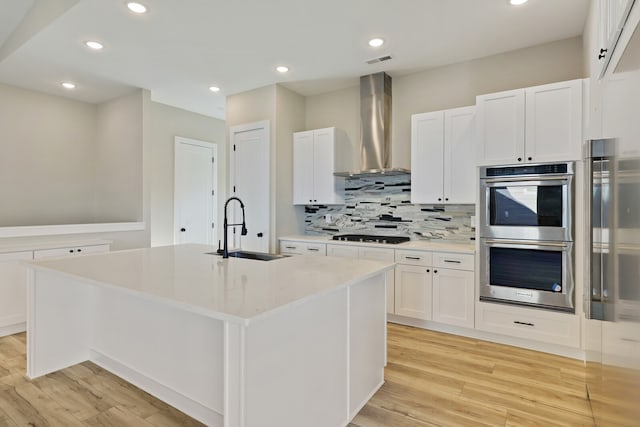 kitchen featuring white cabinets, an island with sink, sink, wall chimney range hood, and double oven